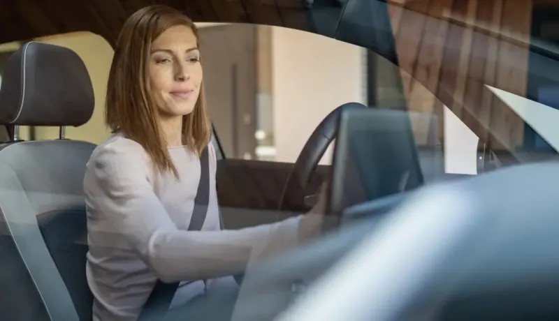 Une femme, souriante, est assise au volant d'une voiture, attachée par sa ceinture de sécurité, en train d'utiliser l'écran tactile de sa voiture.