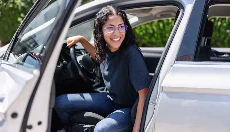 Jeune femme souriante, les cheveux longs, assise sur le siège conducteur d'une voiture blanche.