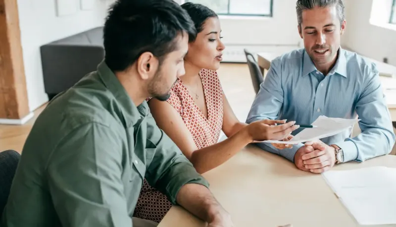 Trois personnes autour d'une table, discutant de documents, probablement lors d'une réunion professionnelle.