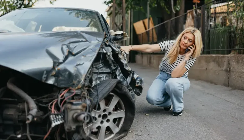 Une femme blonde parle au téléphone, accroupie à côté d'une voiture gravement accidentée.