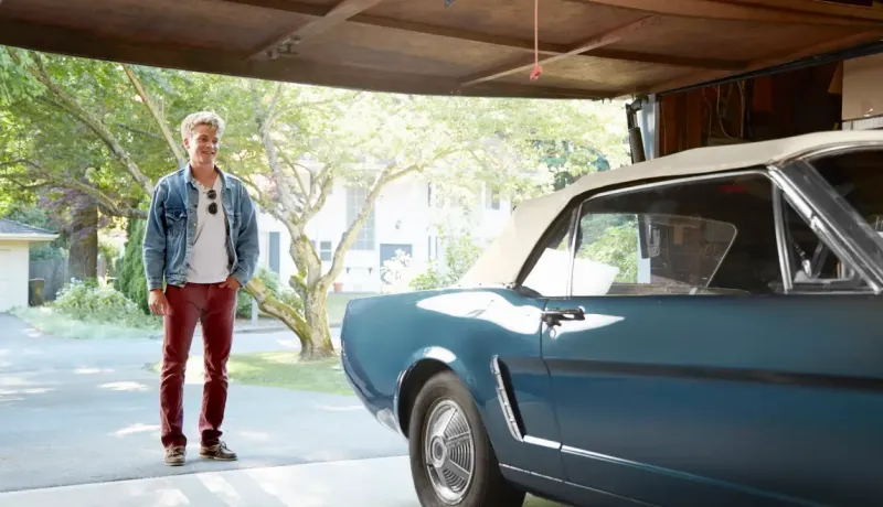 Un jeune homme aux cheveux blonds clairs se tient à côté d'une voiture décapotable bleue vintage dans un garage.