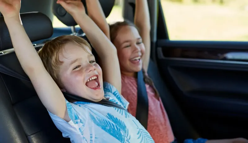 Deux enfants assis à l'arrière d'une voiture, souriant et levant les bras.