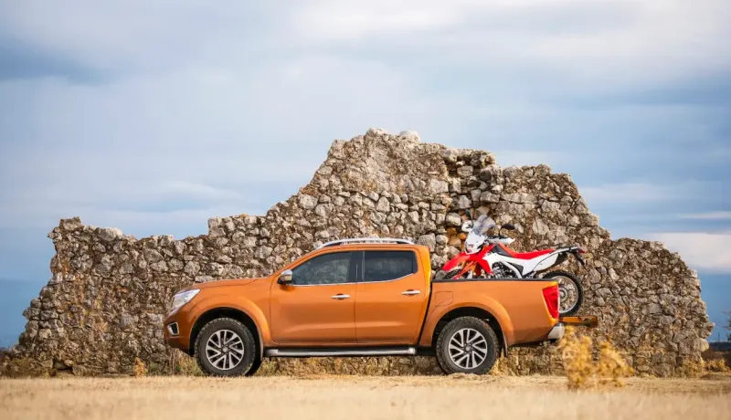Un pickup orange garé devant un ancien mur en pierre avec une moto de cross dans la benne.