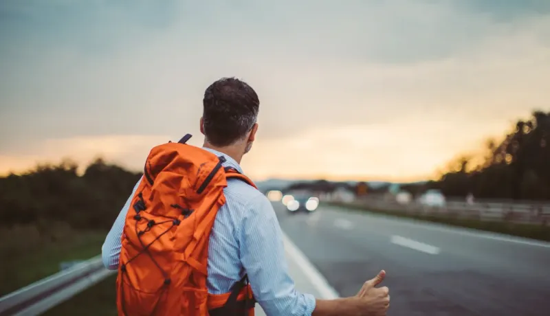 Un homme avec un sac à dos orange fait de l'auto-stop au bord d'une route au coucher du soleil, dos à la caméra.
