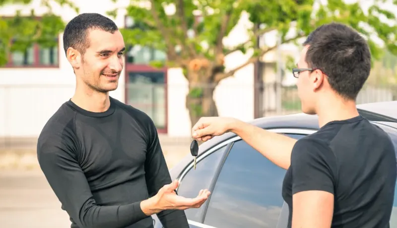 Un homme aux cheveux foncés reçoit les clés d'une voiture d'un autre homme souriant, près d'une voiture.
