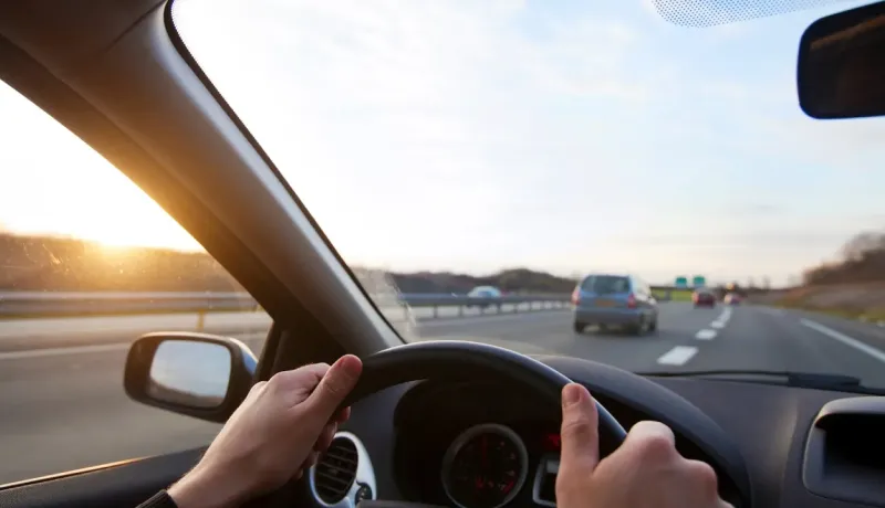 Vue du conducteur sur une autoroute au coucher du soleil, montrant les mains sur le volant et d'autres voitures en arrière-plan.