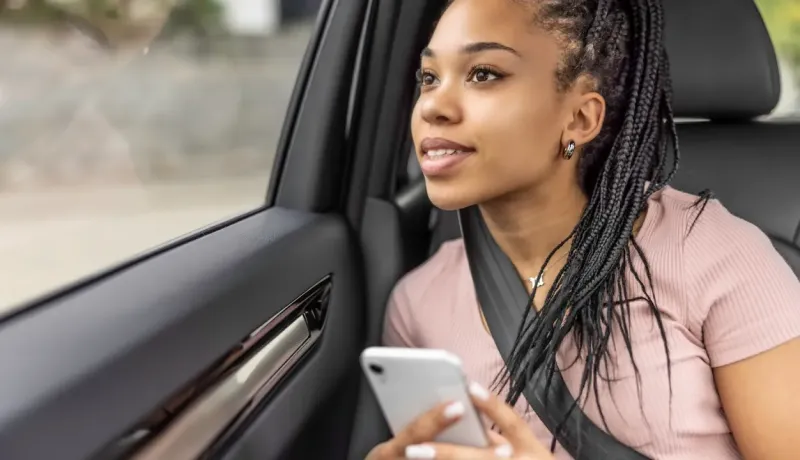 Une jeune femme assise à l'arrière d'une voiture, regardant par la fenêtre son téléphone dans les mains.