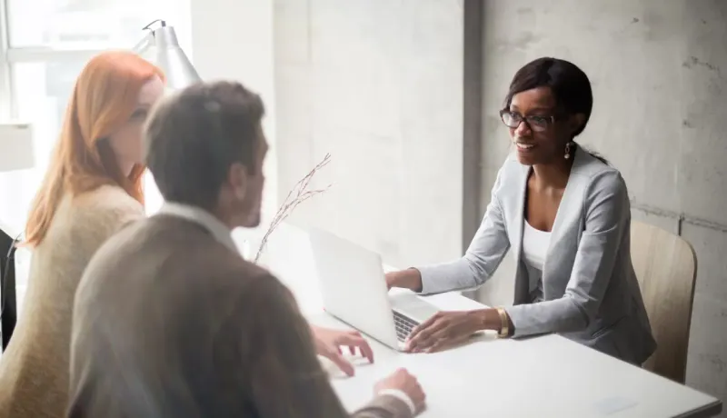 Une conseillère souriante en lunettes discute avec un couple assis à son bureau devant un ordinateur portable.