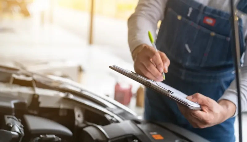 Un mécanicien en salopette écrit sur un presse-papiers, effectuant une inspection sous le capot d'une voiture.