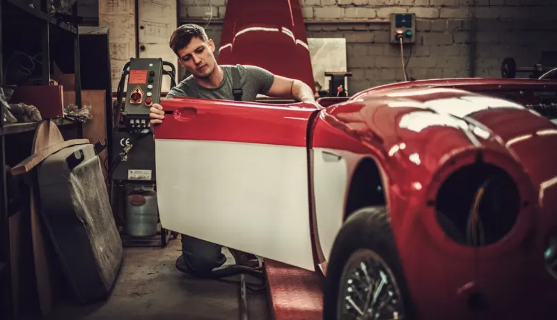Un jeune mécanicien s'affaire sur une voiture de collection rouge et blanche dans un garage, la portière ouverte.