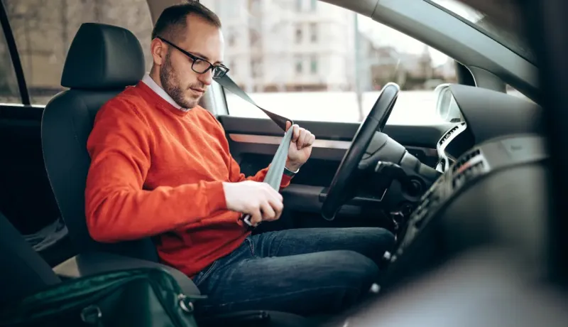Un homme en pull orange et lunettes attache sa ceinture de sécurité dans une voiture, se préparant à conduire.