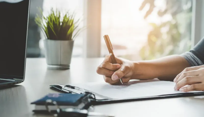 Une personne signe un document avec un stylo sur un bureau, à côté d'un ordinateur portable et d'une plante verte.