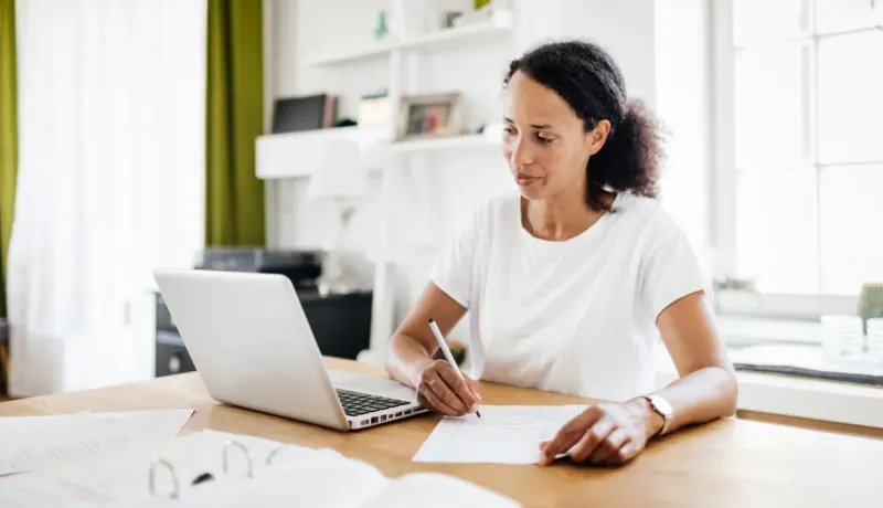 Une femme assise à domicile, écrivant sur un papier et regardant son ordinateur portable.