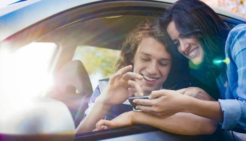 Un jeune couple souriant regarde un smartphone à travers la fenêtre ouverte d'une voiture, le soleil brillant.