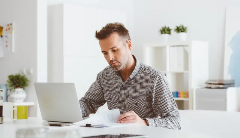 Un homme concentré travaille sur un ordinateur portable, tenant des documents dans un bureau lumineux et épuré.