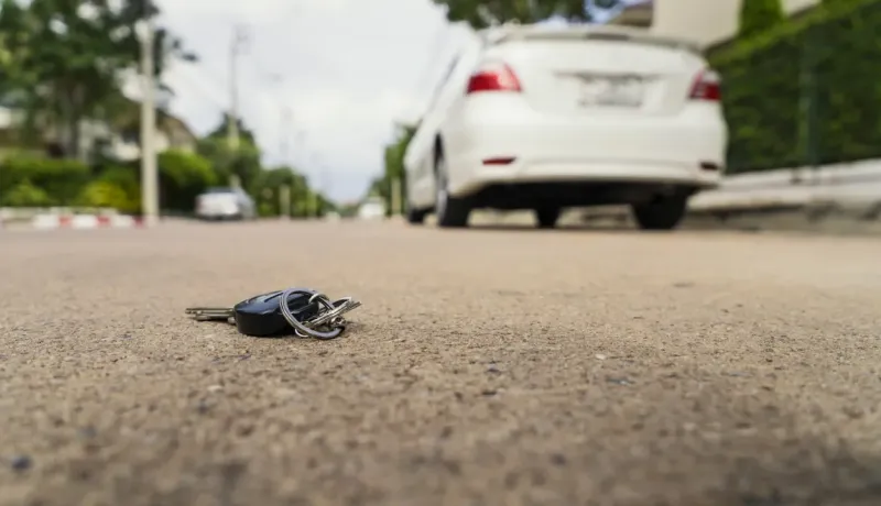 Des clés de voiture noires sont tombées sur la route, avec une voiture blanche garée en arrière-plan.
