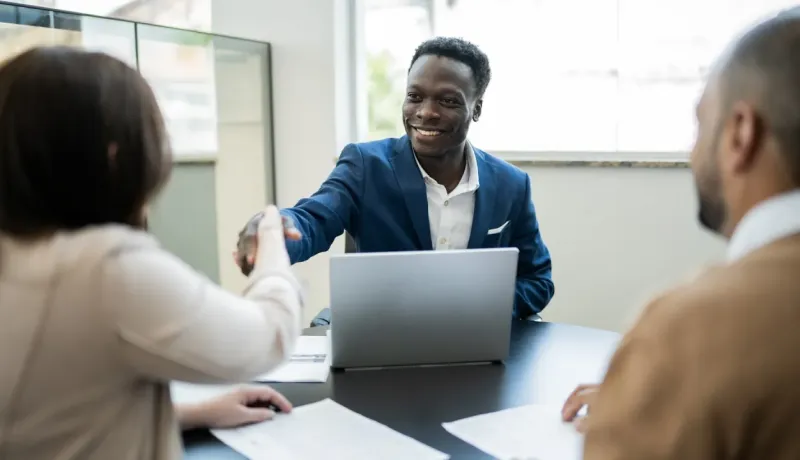 Un homme souriant en costume bleu serre la main d'une femme à un bureau, avec un ordinateur portable ouvert.