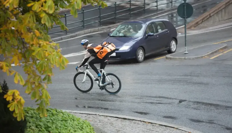 Un cycliste avec un grand sac à dos orange roule sur une route mouillée, avec une voiture en arrière-plan.