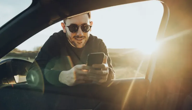 Un homme souriant et portant des lunettes de soleil regarde son téléphone à travers la fenêtre ouverte d'une voiture.