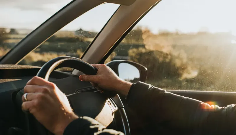Vue de l'intérieur d'une voiture, montrant les mains d'une personne tenant le volant en conduisant au coucher du soleil.