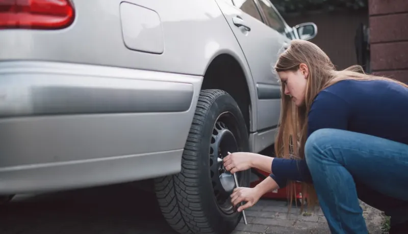 Une femme est accroupie à côté d'une voiture grise, en train de changer une roue ou de serrer les écrous.