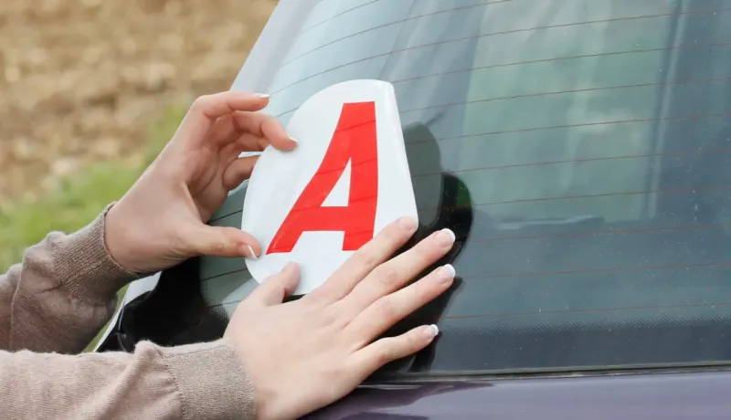 Des mains appliquent un autocollant "A" rouge pour jeune conducteur sur la lunette arrière d'une voiture.