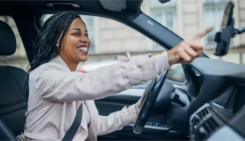 Une femme souriante aux tresses conduit une voiture et utilise son smartphone.