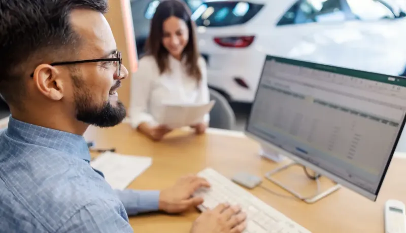 Un vendeur et une cliente discutent, le vendeur utilisant un ordinateur, dans une concession automobile.