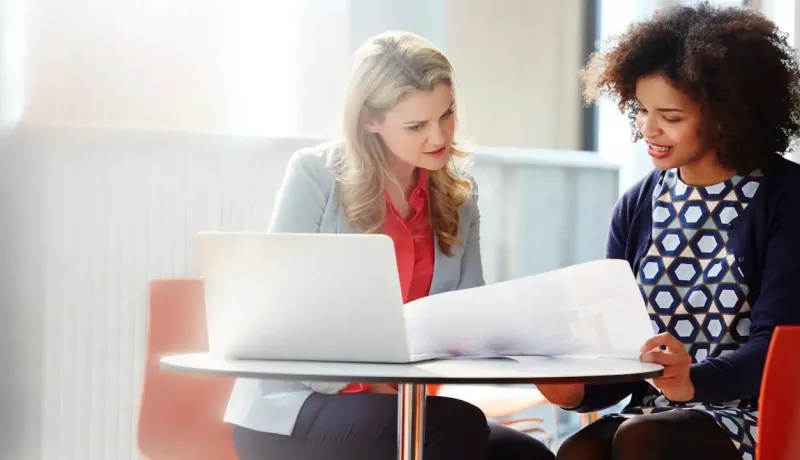 Deux femmes discutent et examinent des documents devant un ordinateur portable sur une table.