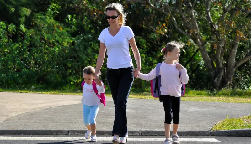 Une femme tient la main de deux jeunes enfants avec des sacs à dos, traversant la route sur un passage clouté.