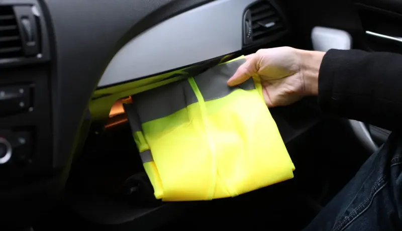 Une main retire un gilet de sécurité jaune fluorescent du vide-poche d'une voiture.