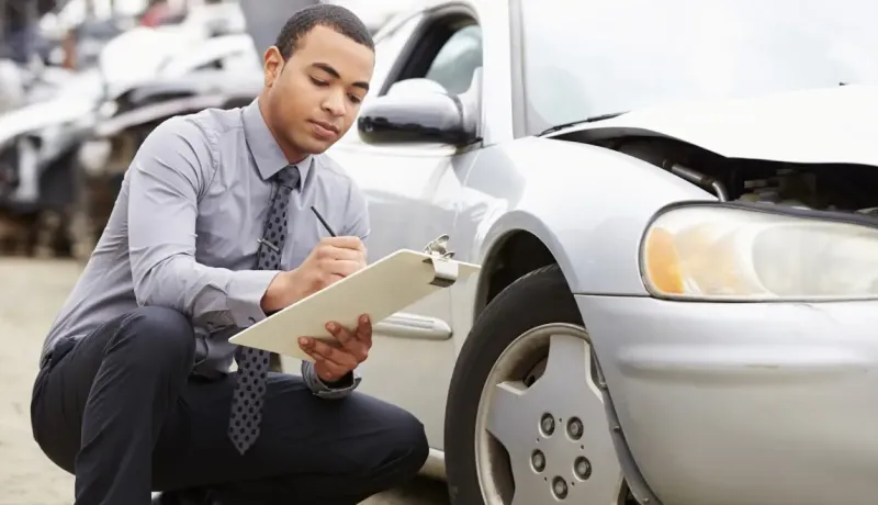 Un expert en sinistres examine une voiture endommagée et prend des notes sur un presse-papiers.