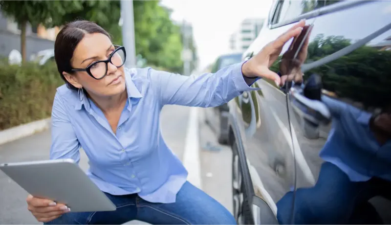 Une femme en chemise bleue et jean examine la carrosserie d'une voiture avec une tablette.