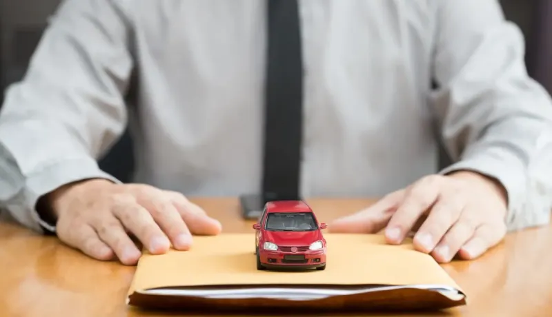 Les mains d'une personne derrière un petit modèle de voiture rouge posé sur une enveloppe marron.