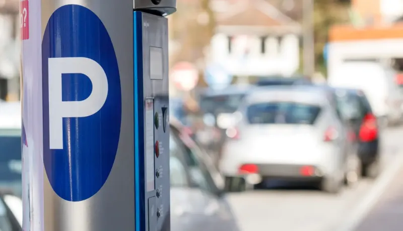 Un horodateur bleu et blanc avec un grand "P" en gros plan, et des voitures garées en arrière-plan.