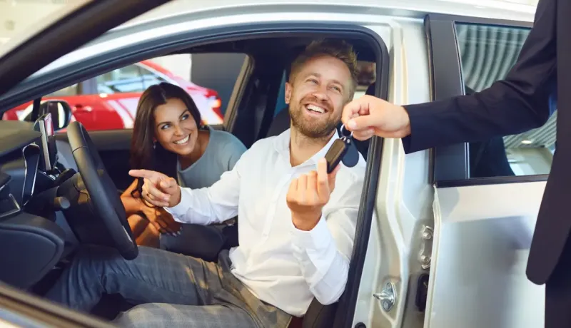 Un homme heureux reçoit des clés de voiture d'un vendeur, avec une femme souriante à ses côtés.
