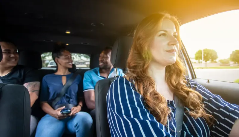 Une femme souriante, au volant d'une voiture, en covoiturage avec trois autres personnes.