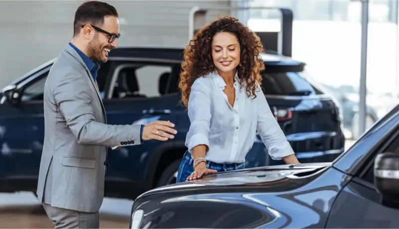 Un vendeur sourit à une femme aux cheveux bouclés qui regarde le capot d'une voiture.