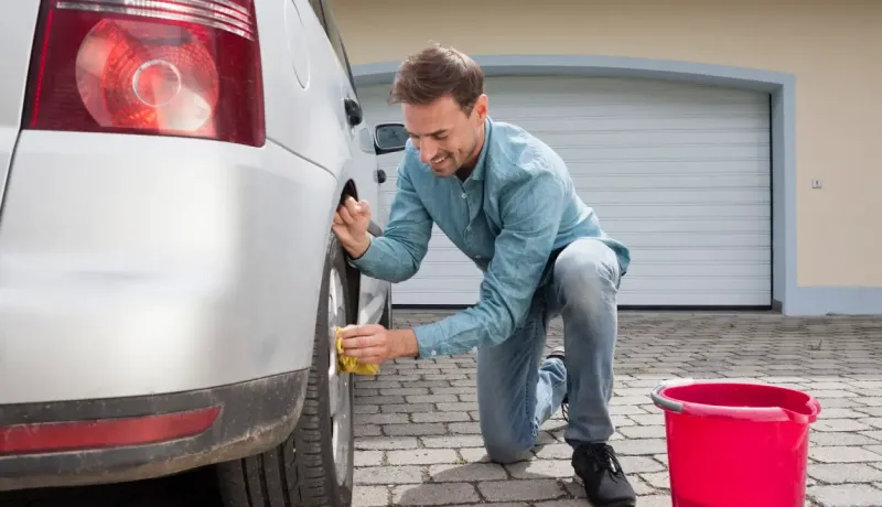 Un homme souriant nettoie la roue arrière d'une voiture grise devant un garage.