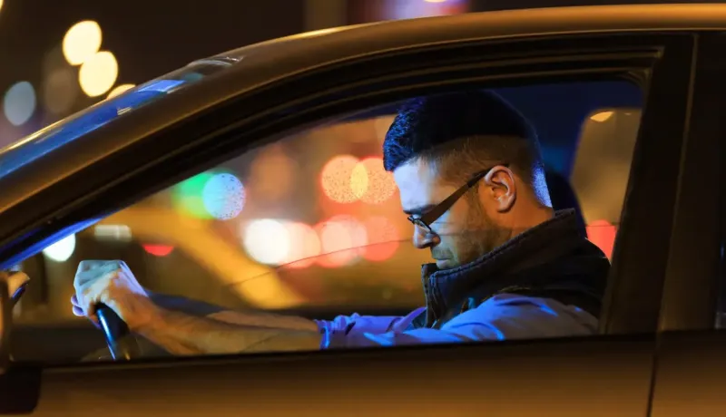 Un homme l'air fatigué au volant d'une voiture la nuit, avec des lumières urbaines floues.