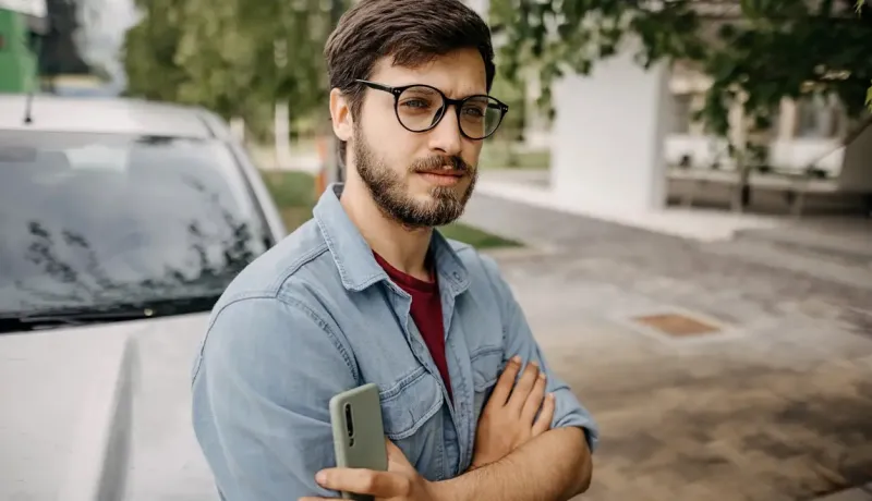 Un homme se tient devant sa voiture avec son téléphone, sur un fond urbain.
