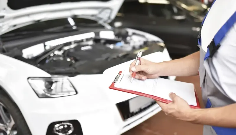 Un mécanicien en salopette prend des notes sur un presse-papiers devant un moteur de voiture ouvert.