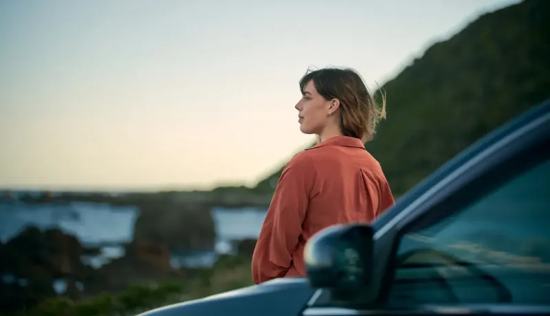 Une femme regardant l'océan à côté de sa voiture.