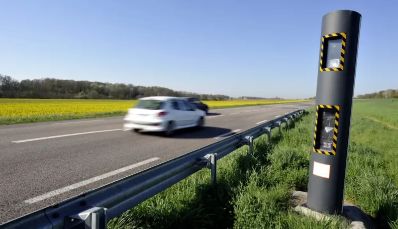 Un radar automatique noir au bord d'une route avec une voiture blanche en mouvement.