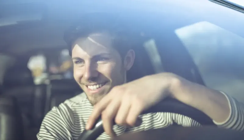 Un jeune homme souriant au volant d'une voiture, regardant à travers le pare-brise.