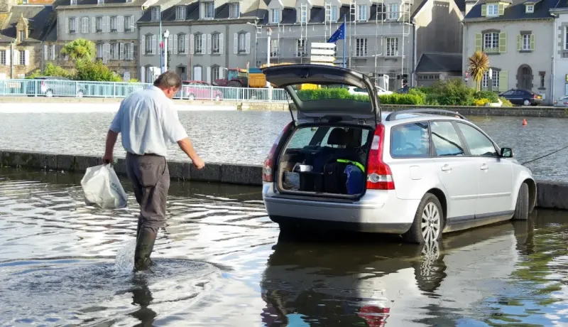Un homme en bottes se tient dans l'eau à côté d'un break gris partiellement immergé.