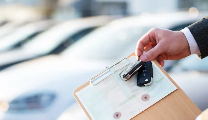 Une main tient des clés de voiture et un presse-papiers avec des documents, devant des voitures neuves.