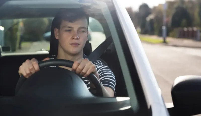 Un adolescent conduit une voiture sans permis, regardant dans son rétroviseur avec une expression concentrée.