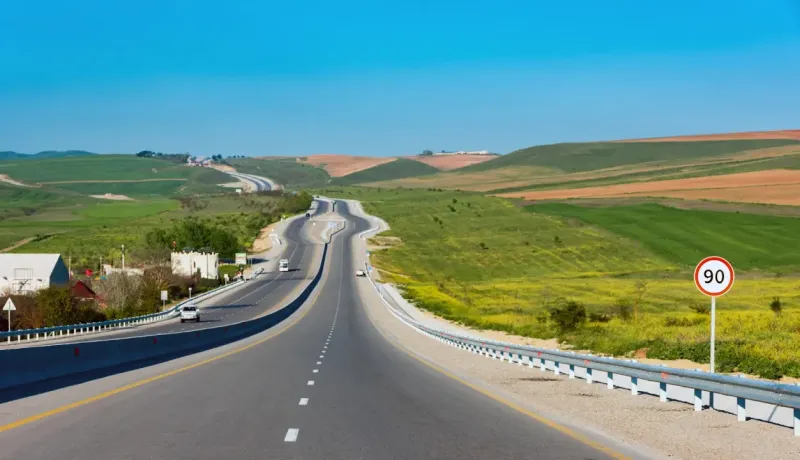 Longue route avec des collines verdoyantes et un panneau de limitation à 90 km/h.