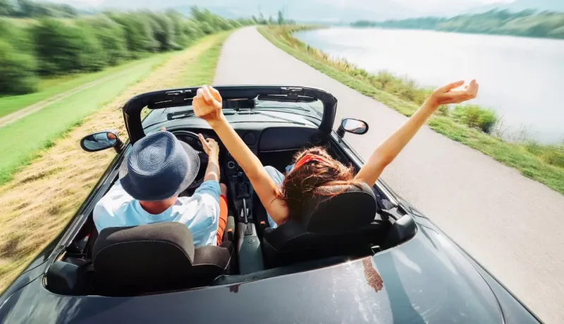Un couple joyeux sur la route dans une décapotable, les bras levés, longeant un lac.